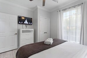 Ein Schlafzimmer mit einem Bett, einem an der Wand montierten Fernseher, einem weißen Kleiderschrank und einem Fenster mit Vorhängen.