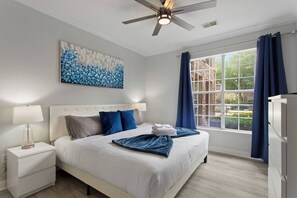 A hotel room with a bed, nightstands, a ceiling fan, a window with curtains, and a painting on the wall. - Royale Blue | 3 BDM | Sleeps 8 | Disney | Resort (Davenport)