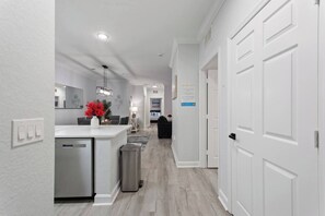 A modern kitchen with a central island, a dining area with a gray sofa, and a hallway leading to a bedroom. - Gentle Breeze | 3BDM | Sleeps 8 | Disney | Resort (Davenport)