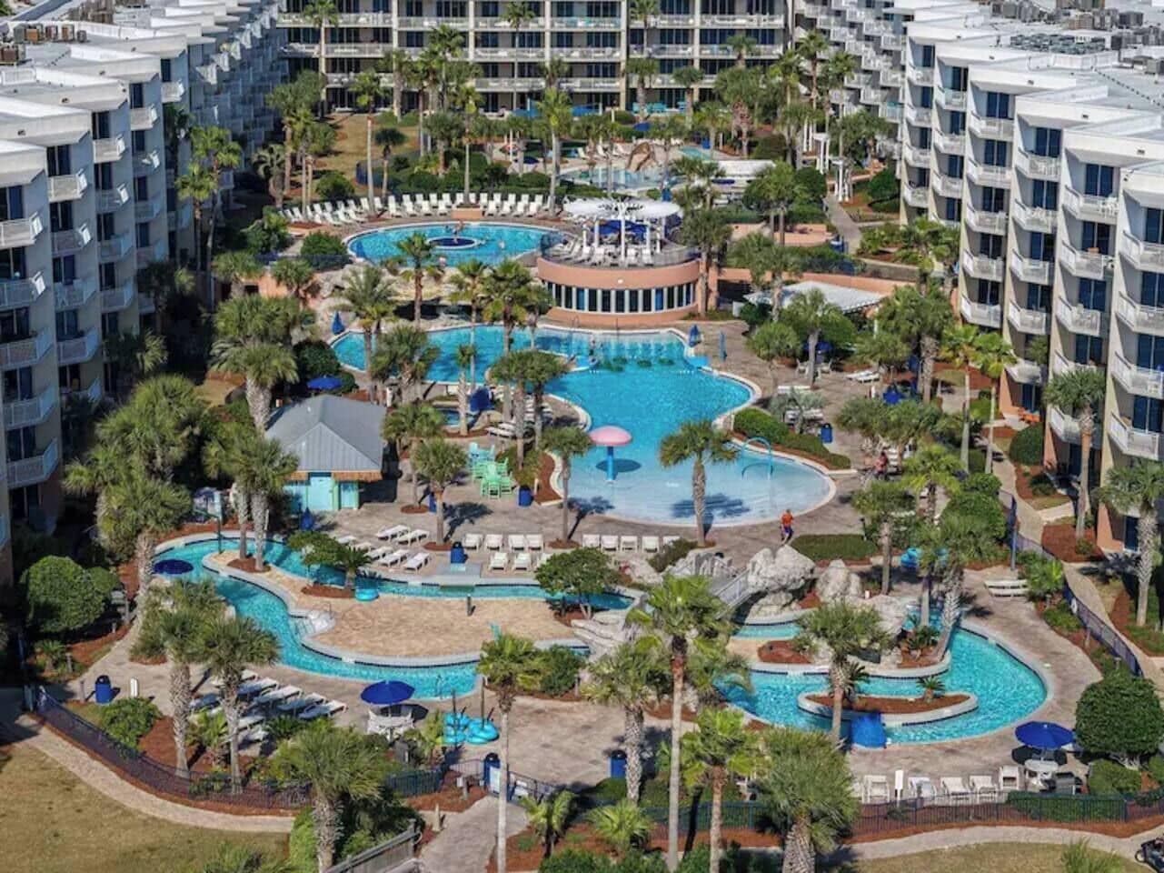 Um hotel com várias varandas, piscina central e palmeiras ao redor.