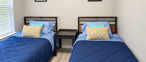 Ein Schlafzimmer mit zwei Betten, jeweils mit blauer Steppdecke und einem Kissen mit der Aufschrift 'hello'. An der Wand befinden sich zwei gerahmte Bilder.