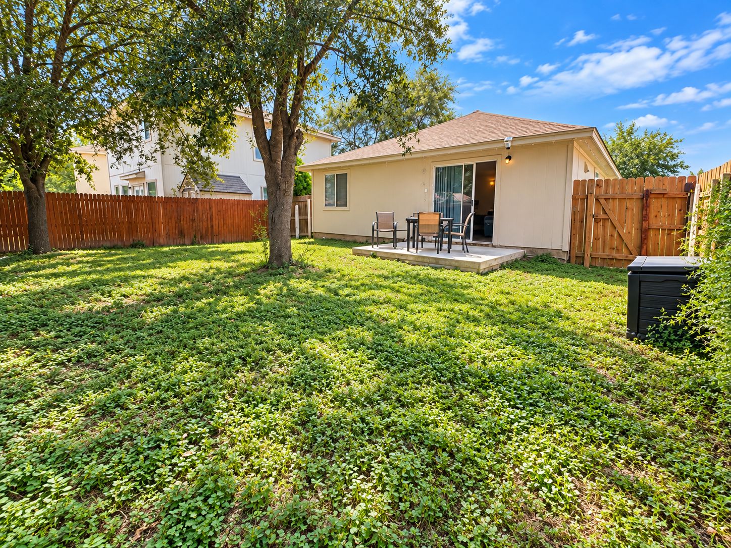 A residential backyard with a house, a wooden fence, a tree, and a patio with outdoor furniture.
