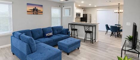 Un salon moderne avec un canapé d’angle, un coin repas avec des chaises, une cuisine avec un bar et un ventilateur de plafond.