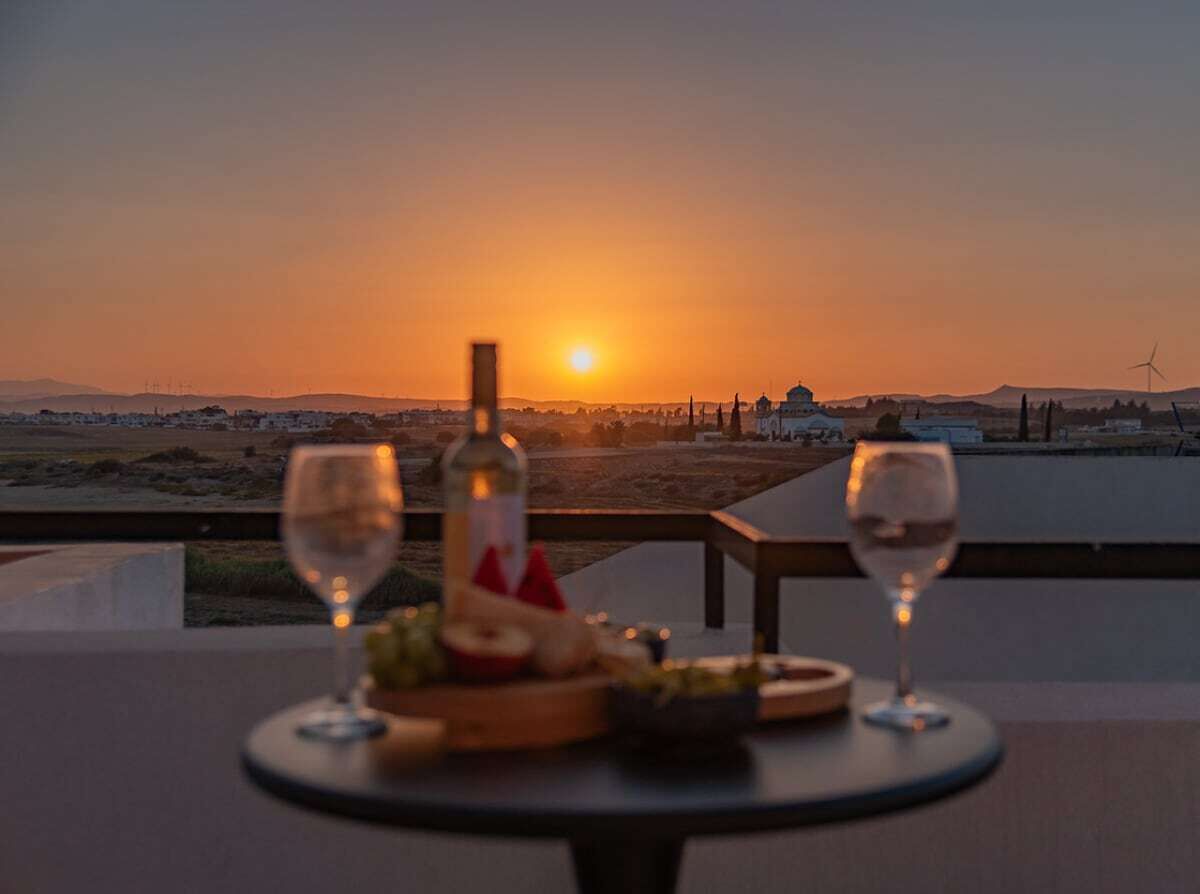 Ein Tisch mit einer Flasche und Gläsern, mit Blick auf einen Sonnenuntergang und eine entfernte Landschaft.
