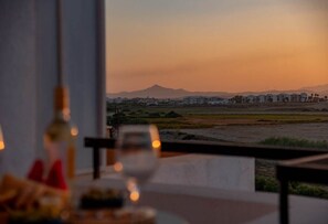 A table with a glass and a bottle, overlooking a landscape with buildings and a mountain. - 3BR Sunset View Retreat Steps from the Beach (Oroklini)