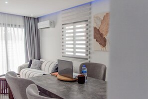 A modern living room with a sofa, a coffee table with a laptop and a water bottle, a dining area with chairs, and a window with blinds. - Modern 2BR Coastal Escape 100m from Sea (Voroklini)