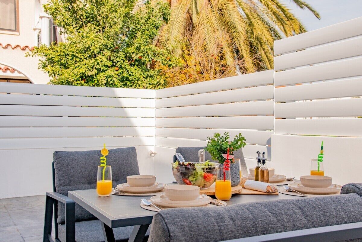 Un espace de restauration en plein air avec une table dressée pour deux personnes, entouré de mobilier blanc à lattes et agrémenté d’un décor luxuriant et verdoyant.