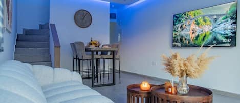 A modern living room with a sofa, a dining area with chairs, a TV on the wall, and a clock.