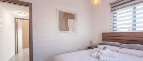 A bedroom with a bed, a bedside table, a hanging light fixture, a window with blinds, and a door leading to another room.