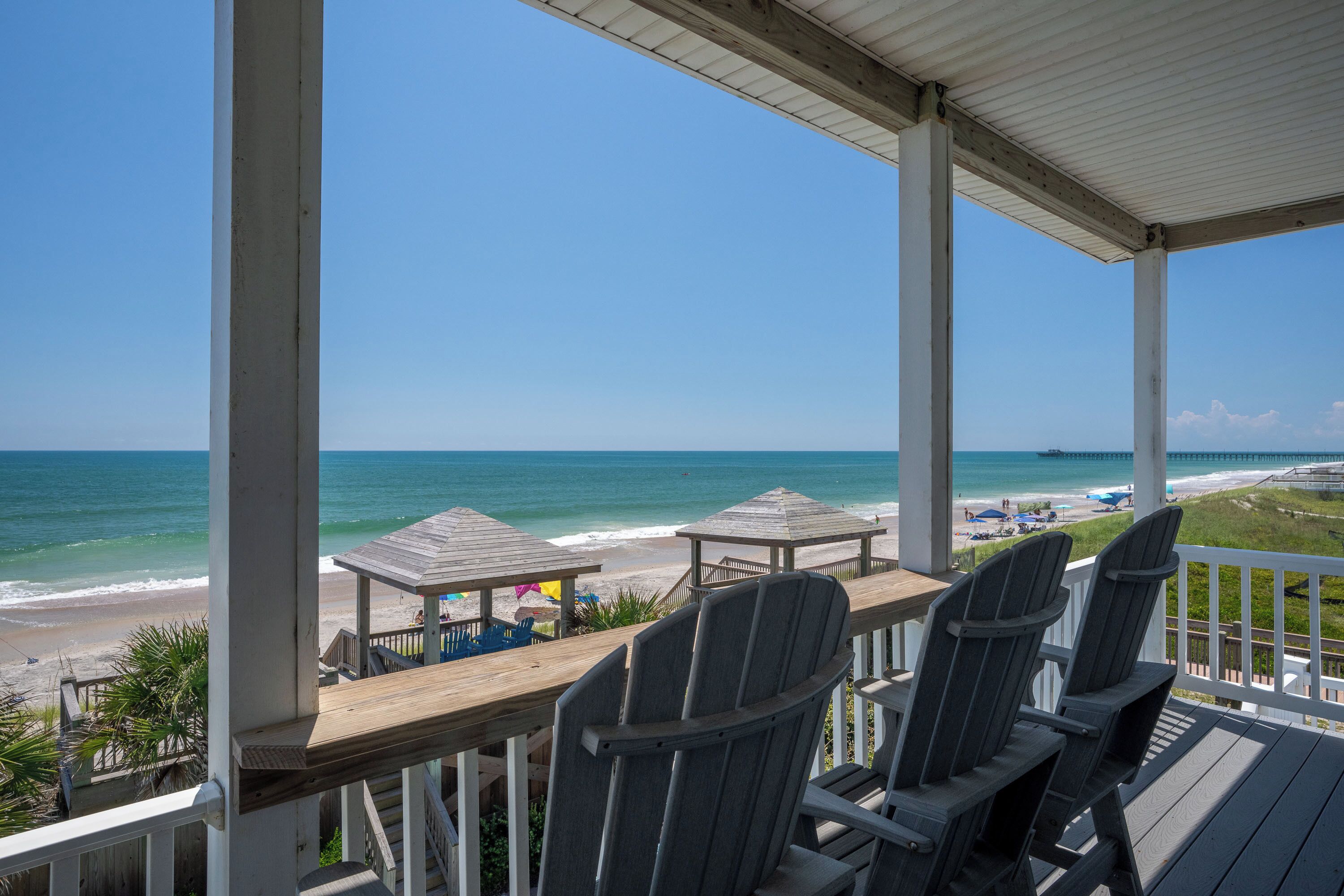 A wooden deck with Adirondack chairs overlooking a beach with a pier.