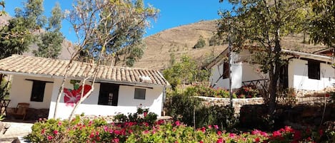 Un edificio bianco tradizionale con tetto in tegole, circondato da piante fiorite e alberi, sullo sfondo di un paesaggio collinare.