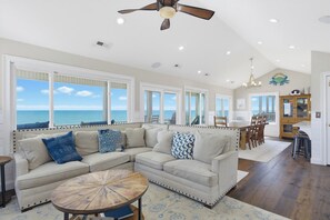 A spacious living room with a large sectional sofa, a round coffee table, and a view of the ocean through large windows. - Oceanfront hot tub and elevator (North Topsail Beach)