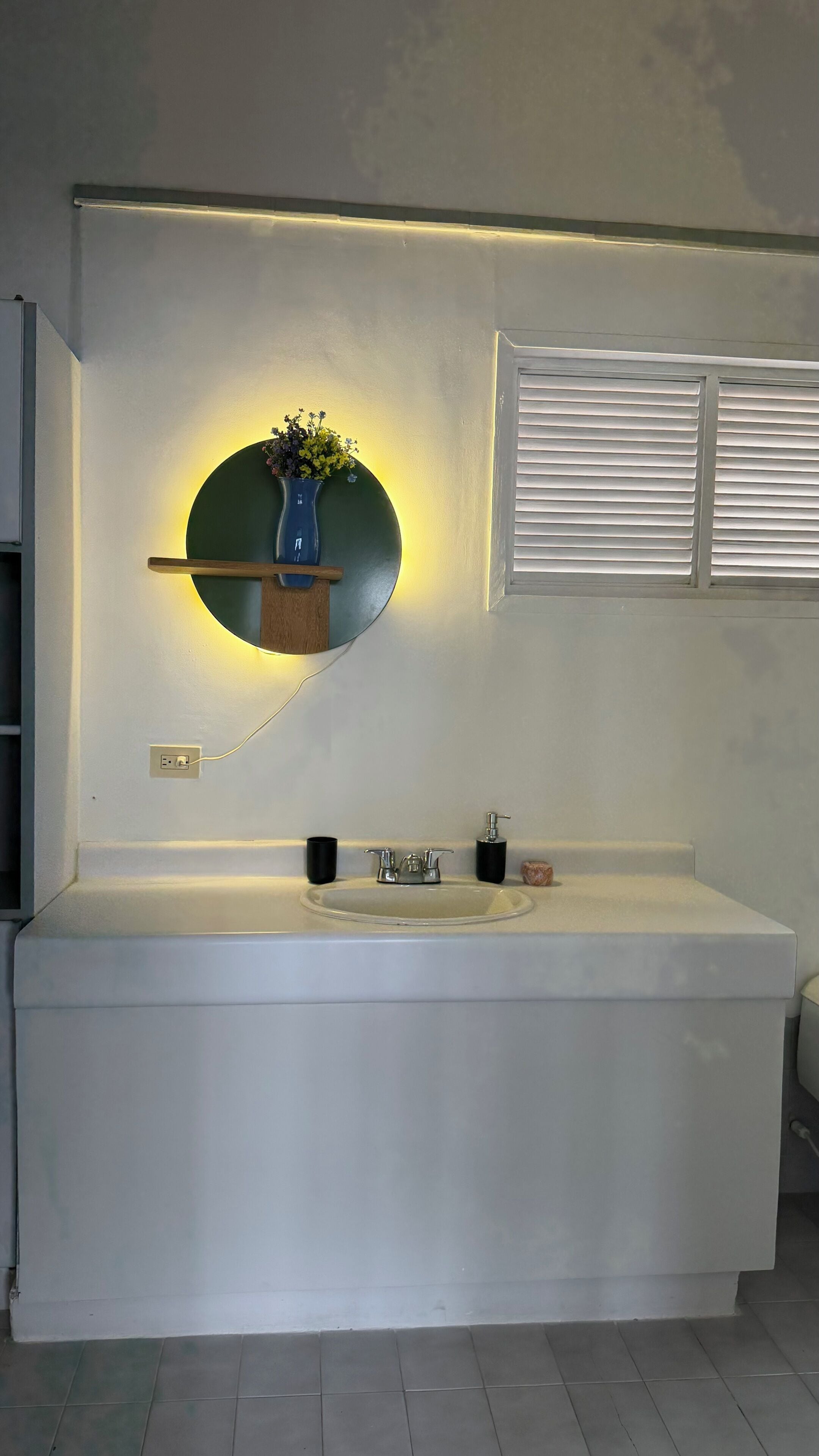 A bathroom with a large white bathtub, a round mirror with a green accent, and a shelf with a vase of flowers.