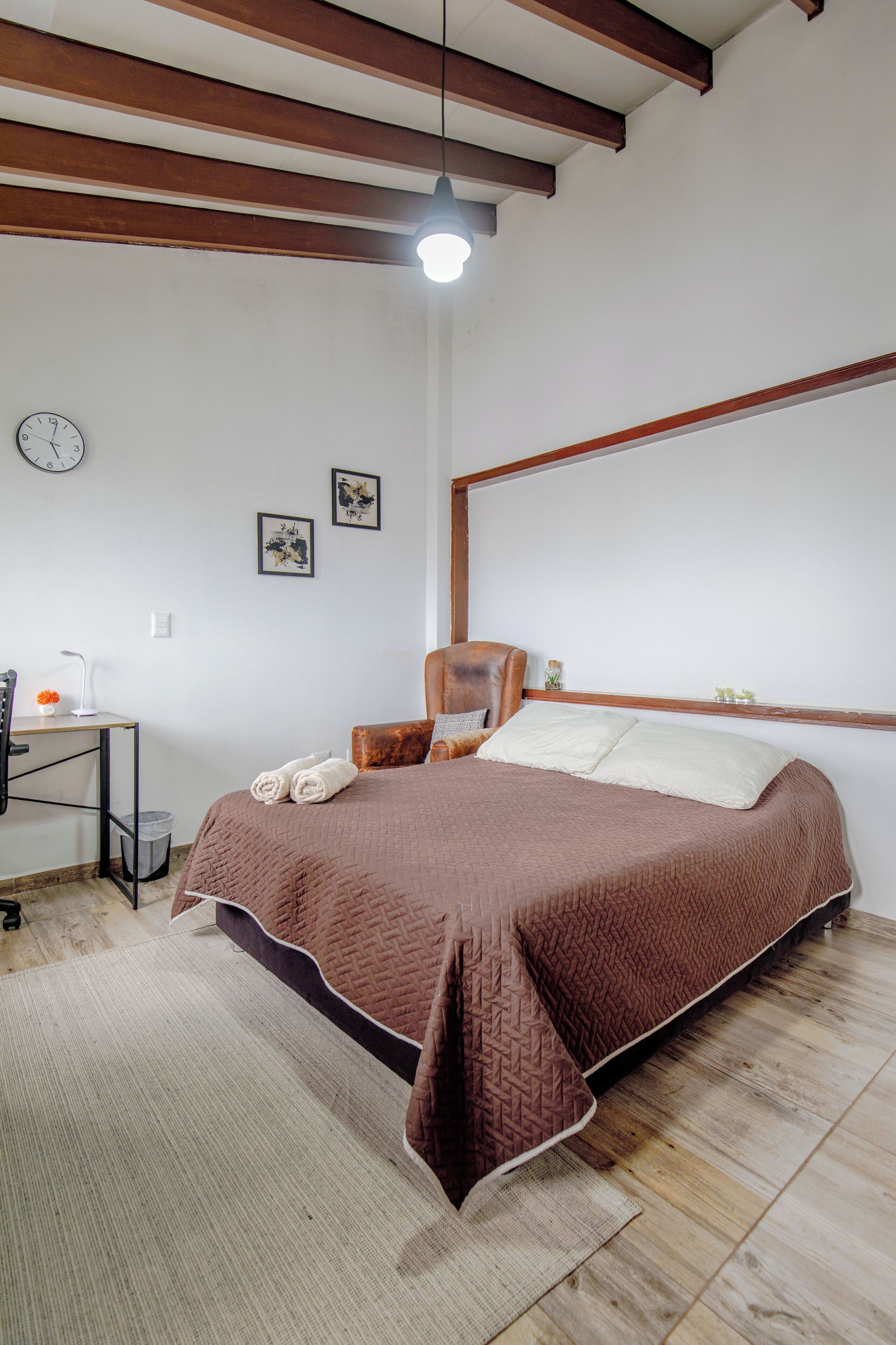 A bedroom with a bed, a desk, a chair, a clock, and two framed pictures on the wall.