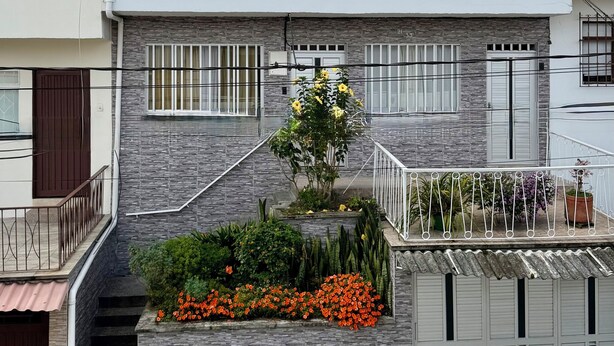 Um edifício de dois andares com escadaria que leva à entrada, um pequeno jardim com flores e um portão de metal.