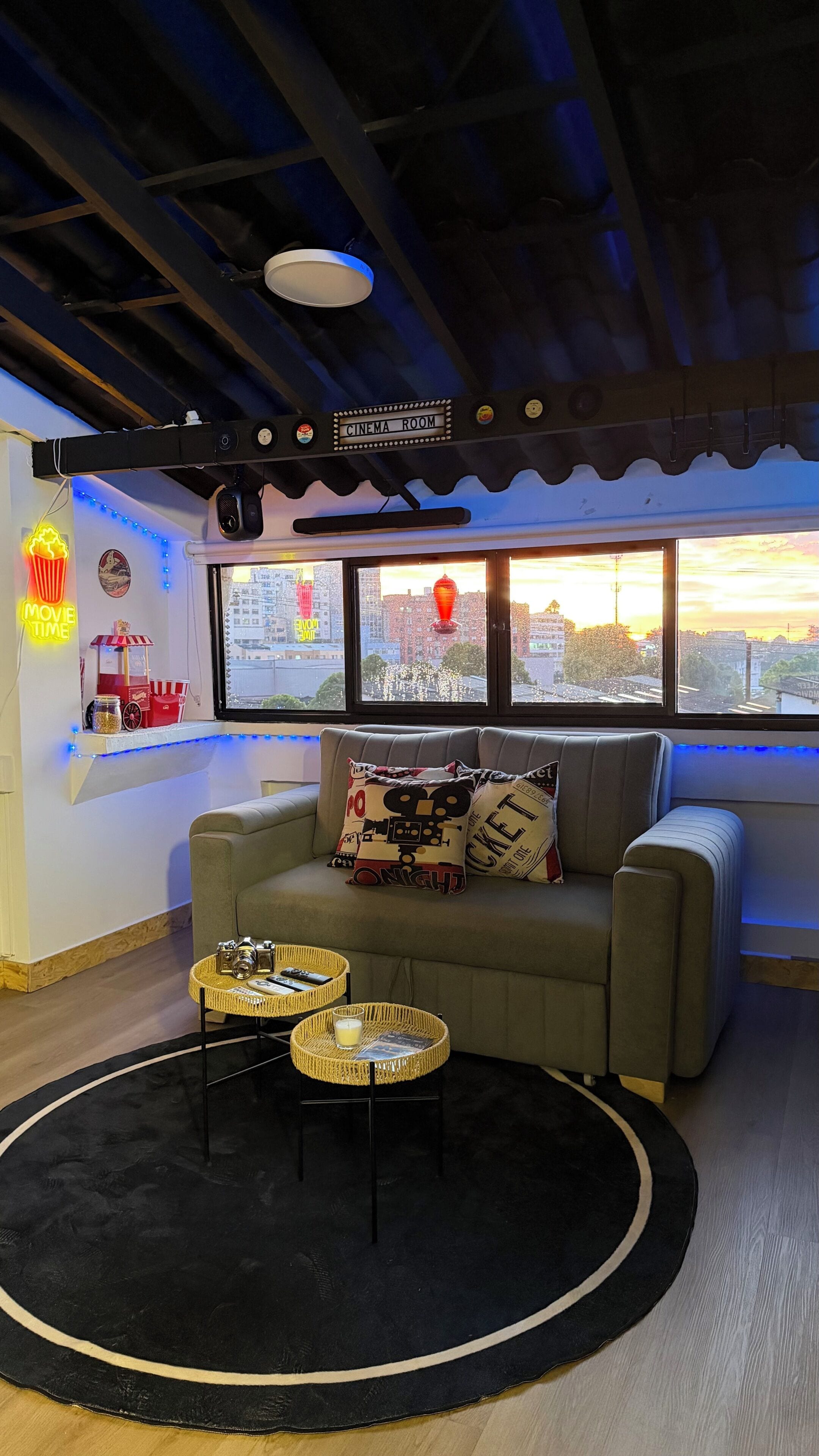 Una sala de cine acogedora con un sofá, dos mesitas y vistas a la ciudad a través de amplias ventanas.