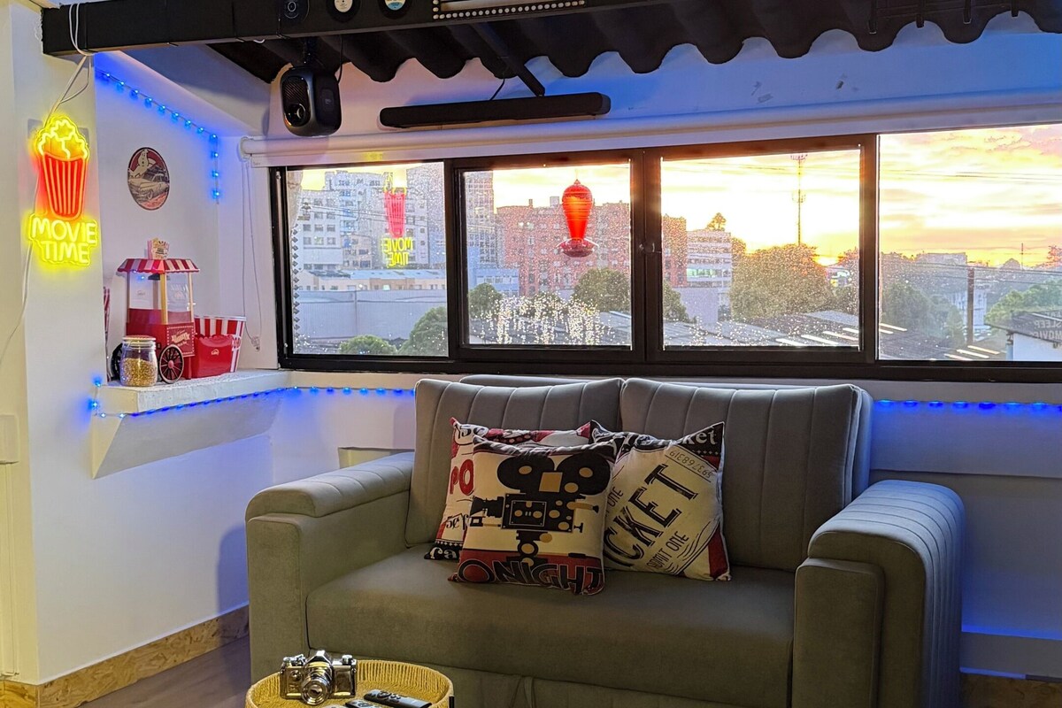 Una sala de cine acogedora con un sofá, dos mesitas y vistas a la ciudad a través de amplias ventanas.