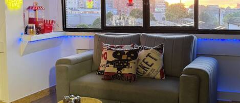 A cozy cinema room with a sofa, two small tables, and a view of the city through large windows.