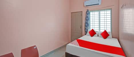 A hotel room with a bed, two chairs, a table with a kettle, and an air conditioning unit.