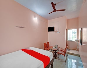 Habitación de hotel con cama, una mesa y sillas, ventilador de techo, televisor montado en la pared y ventana con persianas.
