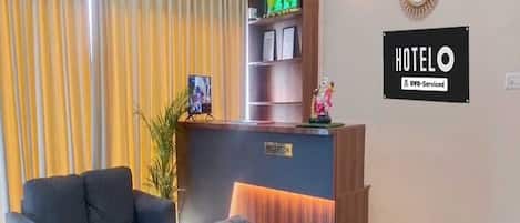 Zona de recepción de un hotel con un mostrador, dos sillones, una planta en maceta y un letrero que indica que el hotel ofrece servicios excesivos.