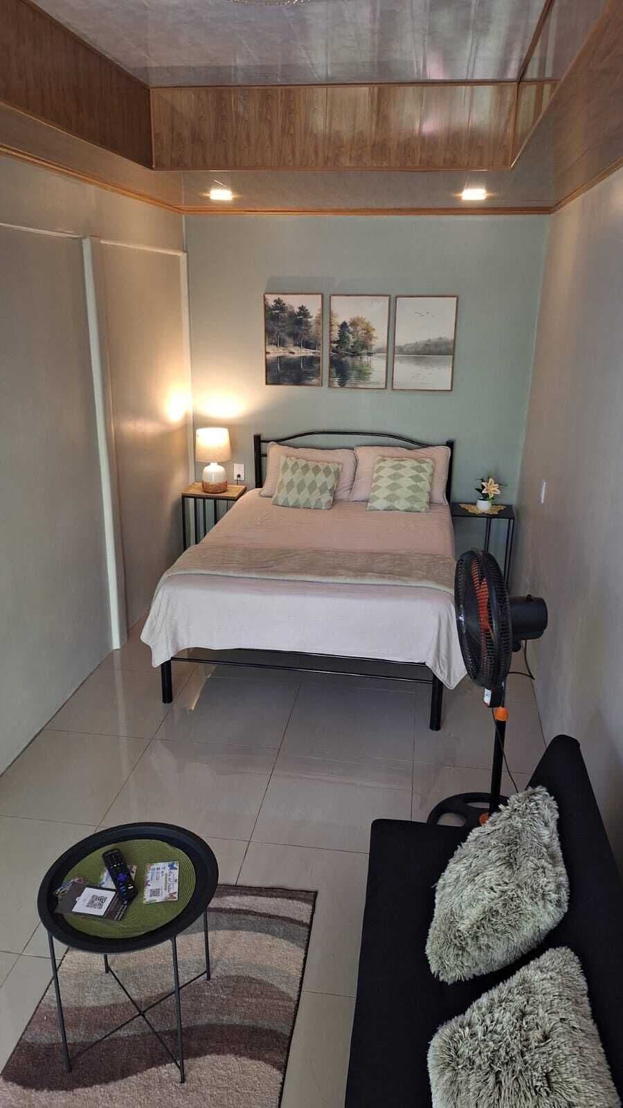 Une chambre à coucher comprenant un lit, des tables de chevet, un ventilateur sur pied, une petite table ronde avec une télécommande et un canapé avec des coussins.