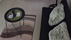 Un dormitorio con cama, mesitas de noche, un ventilador de pie, una pequeña mesa redonda con un control remoto y un sofá con cojines.