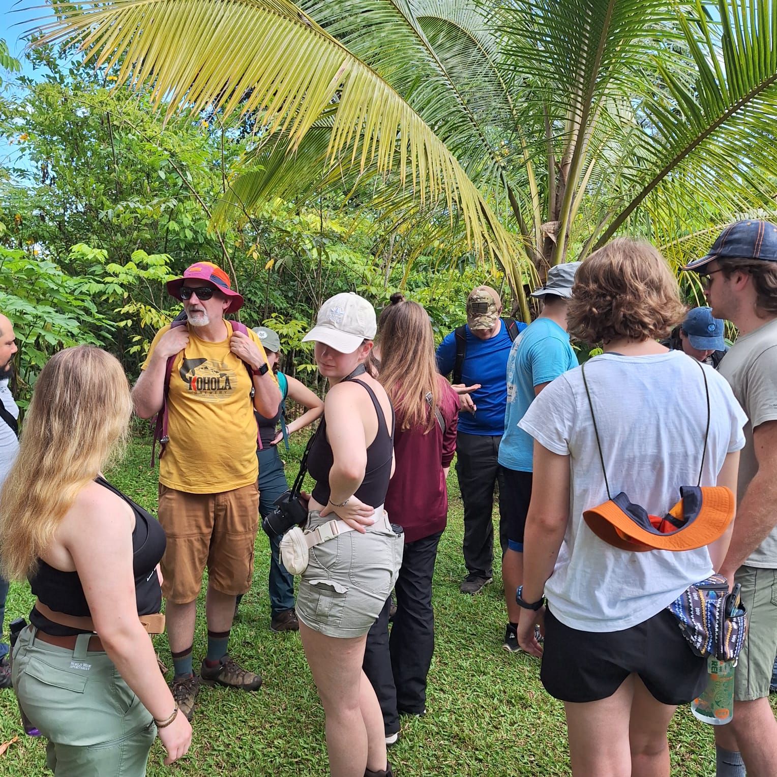 A group of people standing outdoors in a natural setting with greenery and palm trees.