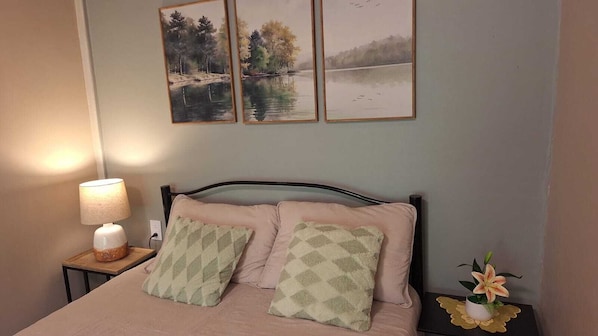 A bedroom with a bed, a bedside lamp, two framed paintings, and a flower vase.