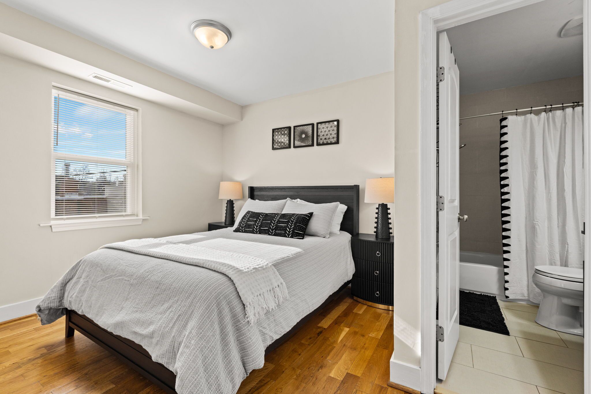 A bedroom with a bed, bedside tables, a window with blinds, and a view of the outside.