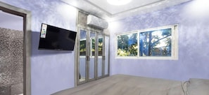 Un dormitorio con cama, televisor montado en la pared, ventana con vistas a árboles y puerta con paneles de vidrio.