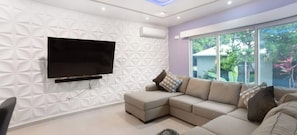 Una sala de estar moderna con un sofá modular, un televisor de pantalla plana montado en una pared con textura y un ventanal con vistas a zonas verdes.