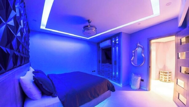 A modern bedroom with a bed, a ceiling light fixture, and a door leading to another room.