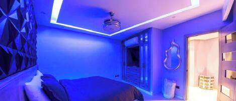 A modern bedroom with a bed, a ceiling light fixture, and a door leading to another room.