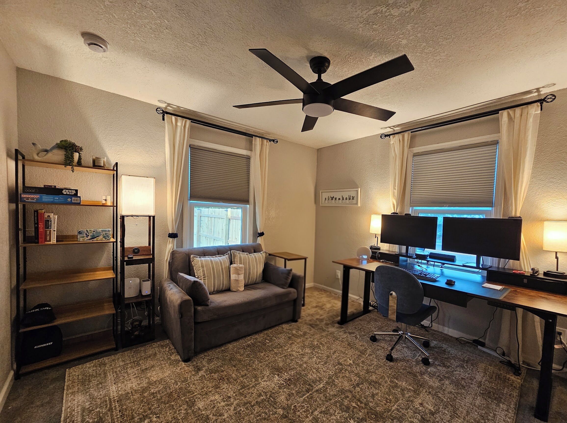 A home office with a desk, two monitors, a chair, a ceiling fan, and a bookshelf.