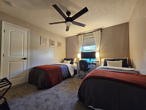 Ein Hotelzimmer mit zwei Betten, einem Schreibtisch, einem Stuhl, einem Deckenventilator und einem Fenster mit Vorhängen.