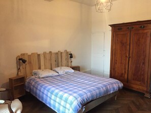 Une chambre à coucher avec une tête de lit en bois, un lit recouvert d’une couverture à carreaux, une table de chevet avec une lampe, une armoire en bois et une porte blanche.