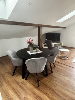 A dining area with a round table, four chairs, a vase with flowers, and a television on a stand.