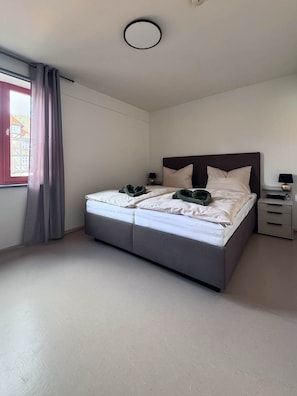 Ein Schlafzimmer mit einem Bett, einem Nachttisch, einer Lampe und einem Fenster mit Vorhängen.