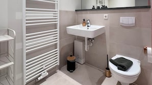 Un baño moderno con lavamanos, espejo y retrete blancos. Hay un perchero y un pequeño cubo de basura.