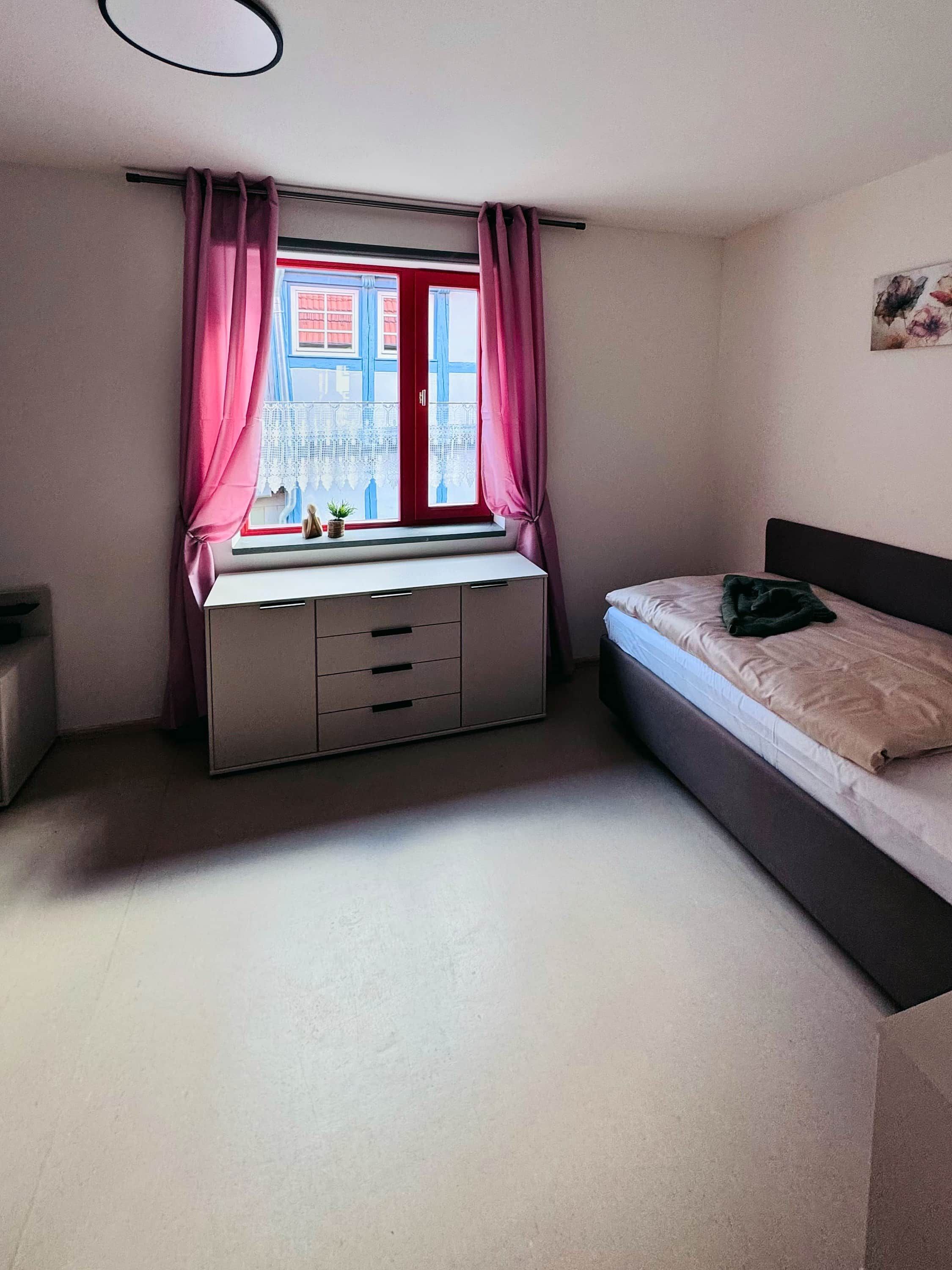 Une chambre à coucher avec un lit, un bureau avec des tiroirs, une fenêtre avec des rideaux et un tableau accroché au mur.