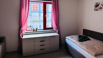 A bedroom with a bed, a desk with drawers, a window with curtains, and a wall-mounted picture.