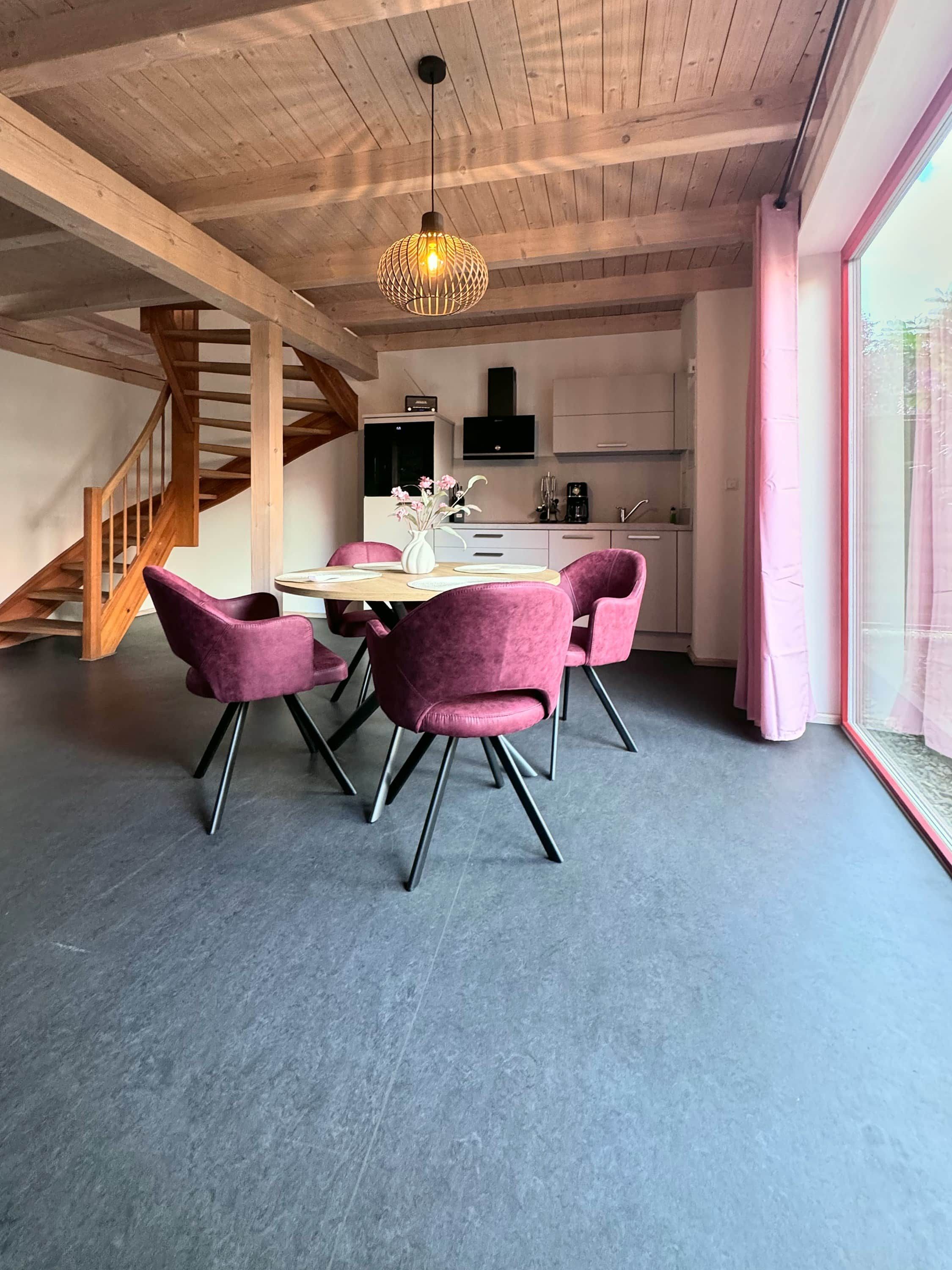 A modern kitchen with a dining area, featuring a staircase, a pendant light, and a large window.