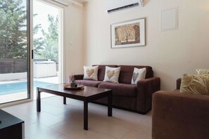 Ein Wohnzimmer mit einer Couch, einem Couchtisch und einem Sessel. An der Wand hängt ein gerahmtes Bild und aus dem Fenster ist ein Blick auf Bäume möglich.