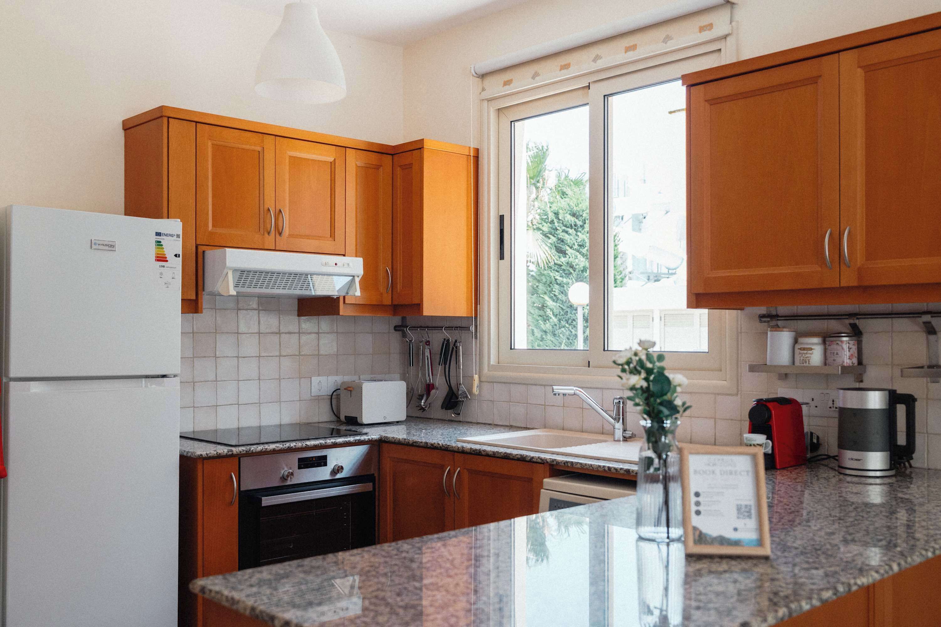 Eine Küche mit Holzschränken, einem weißen Kühlschrank, einem Toaster, einer Kaffeemaschine und einem Fenster mit Blick auf Bäume.