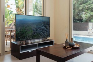 A flat-screen TV on a wooden stand in a room with a large window, a pool, and a dining area with a table and chairs. - 2BR Villa with Private Pool Near Fig Tree Bay (Paralimni)