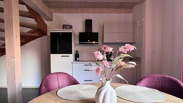 A dining area with a wooden table, two chairs, a vase with flowers, and a kitchen in the background.
