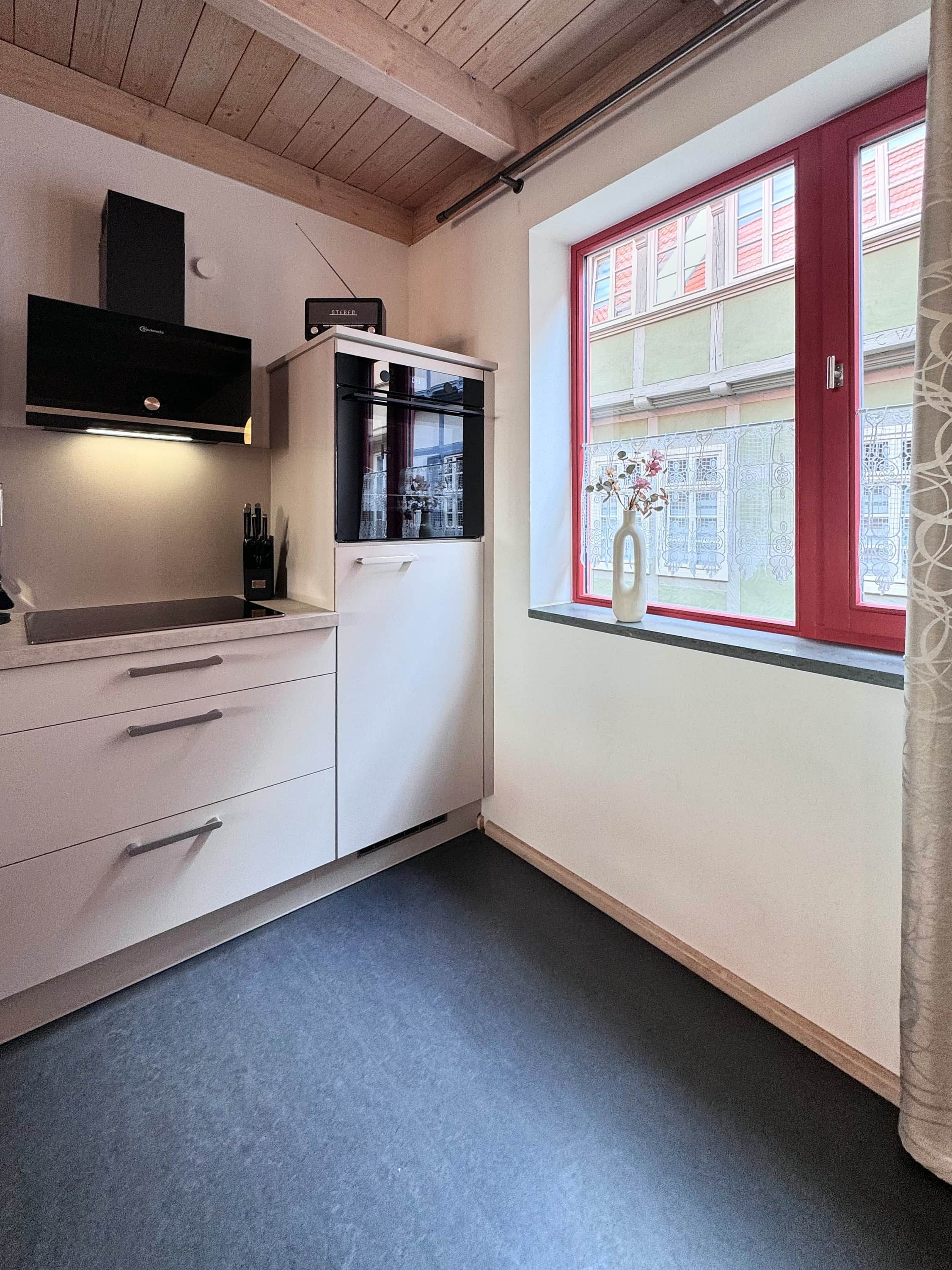 A kitchen with white cabinets, a black range hood, and a window with red trim.