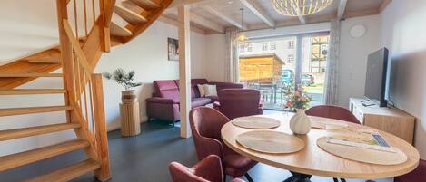 A modern interior with a wooden staircase, a round dining table with red chairs, and a large window with a view of a building outside.