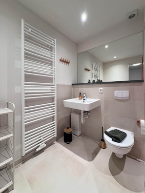 A modern bathroom with a white sink, mirror, and toilet. There is a towel rack and a small trash bin. - Elementa City Lodge Quedlinburg 4 (Quedlinburg)
